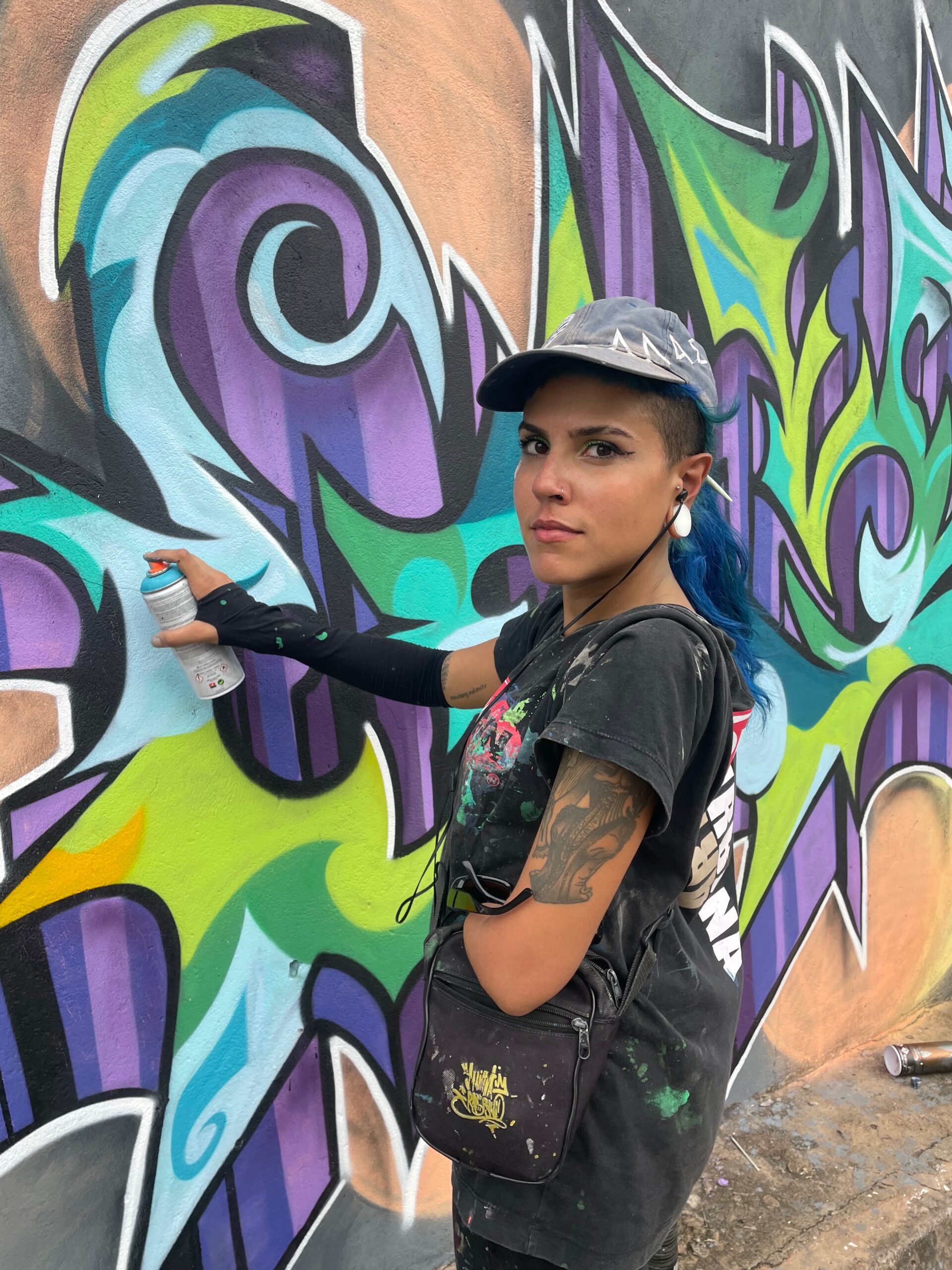 Syner, a graduate in Theory, Criticism and History of Art from the University of Brasília, began graffitiing in November 2019 after voluntarily photographing the 3rd DF Graffiti Meeting. Syner is recognized by the Secretariat of Culture of the Federal District as a cultural agent, number CEAC 10826. Abbreviating the word 'SYNERGY', which means simultaneous action or effort, Syner chose to graffiti organic and pointed letters on the walls of the city, INSPIRED BY the wild style of graffiti. Letters are the origin of the history of graffiti, and graffiti is the propagation of Hip-Hop culture, an element that transforms society. Syner researches letters that suggest rhythm, movement, POINT DIRECTIONS AND SEEK (im)balance. Although characters were her starting point, today she focuses exclusively on writing, an element that has been present in her CONSTANT calligraphy and reading since childhood. Joining the minority of women who are exclusively interested in experimenting with the world of letters in graffiti, in addition to being part of the minority of women with disabilities who do graffiti, Syner seeks to evolve her technique with spray paint and reverberate in time and space the contribution of women and people with disabilities in Hip-Hop culture.